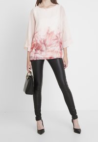 Blusa rosa claro con un diseño floral abstracto en el dobladillo, combinada con leggings de cuero negro brillantes y tacones altos negros. Bolso negro con detalles dorados.