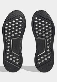 Ett par svarta Adidas skosulor med hexagonalt mönster och synlig vit dämpning under, visade mot en ljus bakgrund.