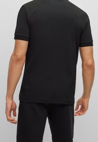 Hombre vestido con una camiseta negra de manga corta y pantalones negros, de pie de espaldas a la cámara frente a un fondo gris claro liso.