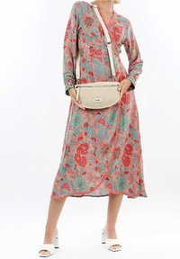 Donna che indossa un abito avvolgente floreale colorato, sandali bianchi con tacco e porta una borsa a tracolla beige con una tasca frontale con zip.