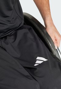 Pantalons de sport noirs avec un logo géométrique blanc, associés à un fauteuil roulant en fibre de carbone, montrant une main saisissant l'accoudoir du fauteuil.