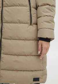 Rūpīgi pildīts beige mētelis ar rāvējslēdzēju priekšā, melnu apdari un mīkstu tekstūru. Iezīmējas ar horizontālām šuvēm un mazu logotipa birku apakšmalā.