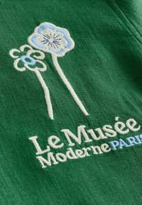 Tissu vert avec deux fleurs stylisées blanches et bleues brodées et le texte "Le Musée Moderne PARIS" en fil blanc et bleu.