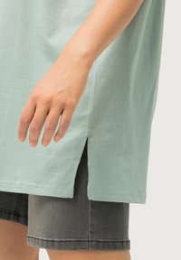 T-shirt di cotone verde chiaro con spacchi laterali, abbinato a pantaloni corti grigio scuro. Enfasi sulla texture e su una vestibilità casual e rilassata.