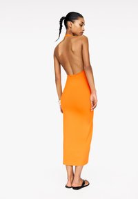 Robe orange sans dos avec une silhouette ajustée, longueur mi-mollet, texture lisse et un col halter. Associée à des sandales noires simples.