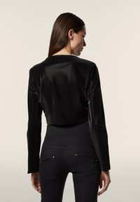 Giacca nera in finta pelle, tagliata, con maniche lunghe; trama liscia e design del collo semplice. Indossata sopra jeans scuri con dettagli hardware sottili.