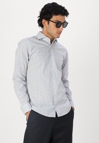 Witte blouse met blauwe bloemenpatroon, spread collar en lange mouwen. Casual gedragen met zwarte zonnebril en donkere broek.