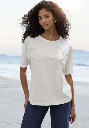 Junge Frau mit lockigem Haar, die ein weißes, mit Blumen besticktes Kurzarmoberteil und dunkle Jeans trägt und an einem ruhigen Strand mit Bergen im Hintergrund steht.