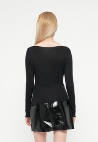 Donna bionda con capelli lisci, che indossa un top nero a maniche lunghe e una gonna nera lucida, rivolta di spalle su uno sfondo bianco.