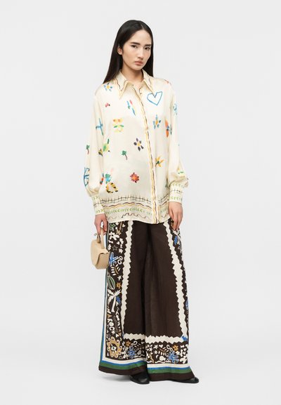 Γυναίκα με μακριά σκούρα μαλλιά, ντυμένη με κρεμ μπλούζα με πολύχρωμα σχέδια doodle και μαύρο παντελόνι με φαρδιά μπατζάκια και floral περιθώρια, κρατώντας μια μπεζ τσάντα.