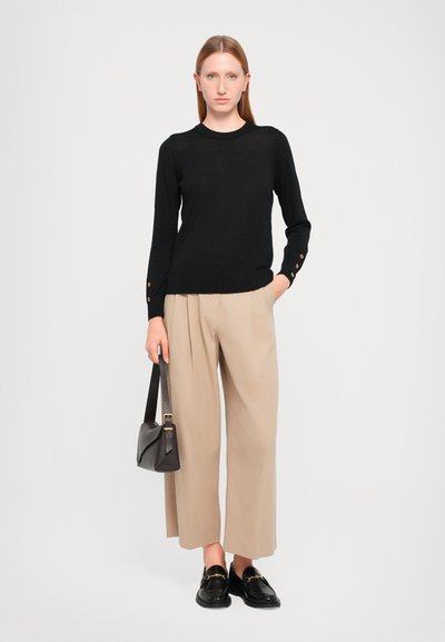 Sort striketrøje, beige brede bukser, sorte loafers med guld accenter, holder en lille mørk håndtaske, glatte stofteksturer.