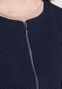 Chaqueta de punto azul marino con textura acolchada, cierre de cremallera suave y cuello redondo, superpuesta sobre una blusa beige.