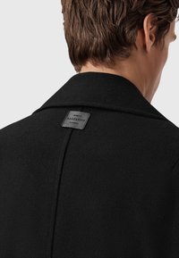 Mustavilla takki, jossa on räätälöity muotoilu, korkea kaulus ja takana korttilappu, jossa lukee "AllSaints London" kohokuvioituna.