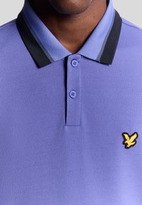 Primo piano di una persona che indossa una polo viola con un colletto a righe nere e viola chiaro e un logo di un uccello giallo sul petto.