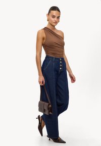 Haut marron à une épaule avec détail froncé, pantalon en denim bleu taille haute avec accents de boutons dorés, et petit sac à main marron.