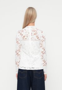 Top in pizzo bianco con maniche lunghe, motivo floreale, scollatura a onde e schiena trasparente. Indossato con pantaloni in denim scuri a vita alta.