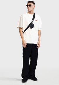 T-shirt in cotone color crema con testo grafico rosso, abbinata a pantaloni in velluto a coste neri e una piccola borsa a tracolla nera. Accessori comprendono occhiali da sole scuri.