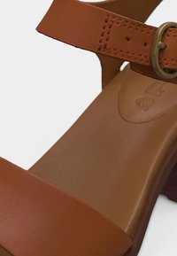 Sandale en cuir marron avec bride ajustable et boucle métallique sur une semelle lisse et un talon bas en bois.