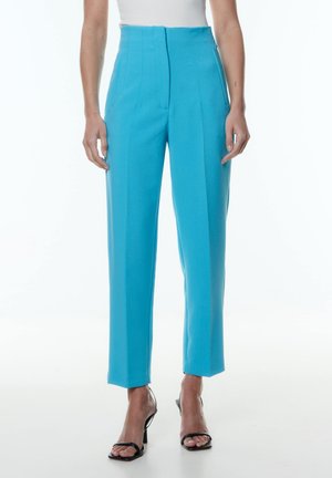 Pantalons cropped taille haute en tissu bleu vif avec une texture lisse, présentant des plis à l'avant et des poches latérales. Associés à des talons noirs.
