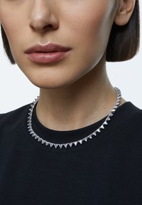 Sølv halskæde med trekantede vedhængsdesigns, der har en skinnende finish. Båret over en sort skjorte, hvilket viser et nærbillede af halsudskæringen.