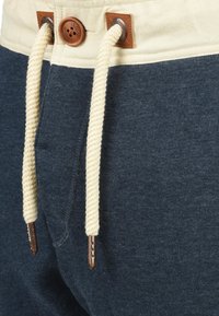 Dark blue sweatpants feature a cream waistband with a drawstring. The drawstring is braided and secured with metallic grommets and a wooden toggle.