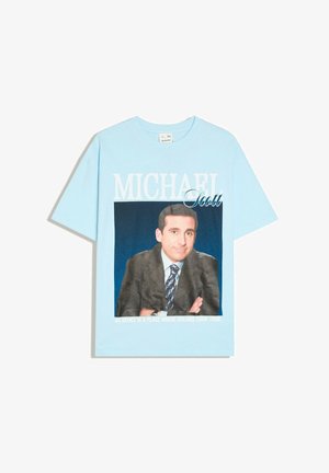 Jasnoniebieska koszulka z zdjęciem mężczyzny w garniturze i krawacie, tekst "MICHAEL Scott" i "An office is a place where dreams come true" poniżej.