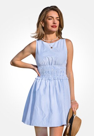 Donna con vestito senza maniche a righe blu e bianche che tiene un cappello di paglia, indossa collane di conchiglie a strati e guarda di lato.