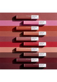 Sept rouges à lèvres liquides Maybelline Super Stay Matte Ink dans différentes teintes, présentant des tubes rectangulaires avec un capuchon blanc, disposés sur des arrière-plans colorés.