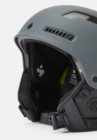Casco de esquí en gris mate con puertos de ventilación, acolchado interior negro, correa ajustable y detalles protectores visibles.