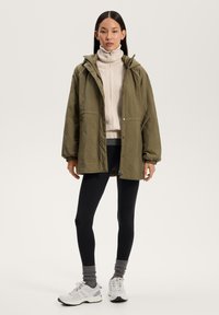 Olivgrønn quiltet jakke med hette, parret med en kremfarget høyhalset genser, svarte leggings og hvite-grå sneakers.