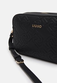 Sort struktureret håndtaske med prægede mønstre, guld logo, lynlås lukning og justerbar rem. Kompakt rektangulær form.