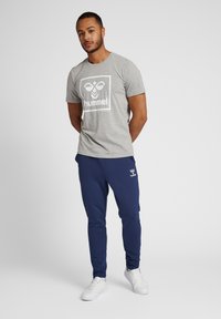 Man står med händerna bakom ryggen, klädd i en grå hummel-t-shirt, marinblå hummel-joggingbyxor och vita sneakers framför en plain grå bakgrund.