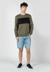 Felpa verde oliva con pannello nero, abbinata a pantaloncini di jeans chiari. La modella indossa scarpe da ginnastica blu navy. Design semplice, vestibilità casual.