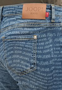 Primer plano de unos vaqueros de mezclilla azul con patrón repetido de texto blanco "JOOP! JEANS", un bolsillo trasero y una etiqueta de cuero marrón con la marca.