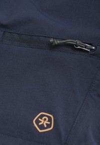 Marinblått tyg med en fin textur, med en dragkedjeficka och en logotyp i orange. Designen inkluderar en dekorerad snodd för justerbarhet.