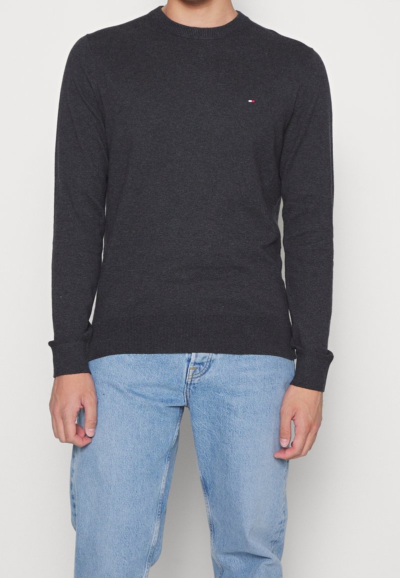 Homme portant un pull à col rond à manches longues gris foncé avec un petit logo sur la poitrine et un jean bleu clair, debout devant un fond blanc.