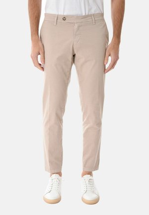 Uomo che indossa pantaloni beige slim fit con chiusura a bottone e cerniera, in piedi con le braccia rilassate, abbinati a sneaker bianche su sfondo bianco.