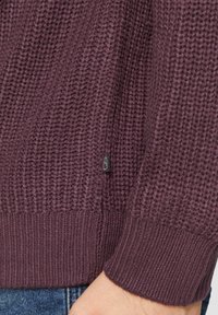 Gehaakte kastanjebruine trui met een textured patroon, geribbelde manchetten en zoom, met een klein zwart logo label aan de zijkant.