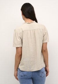 Kvinde med langt, mørkt hår iført bluse med korte ærmer i beige med flæsekant og blå jeans, vist fra ryggen mod en ensfarvet baggrund.