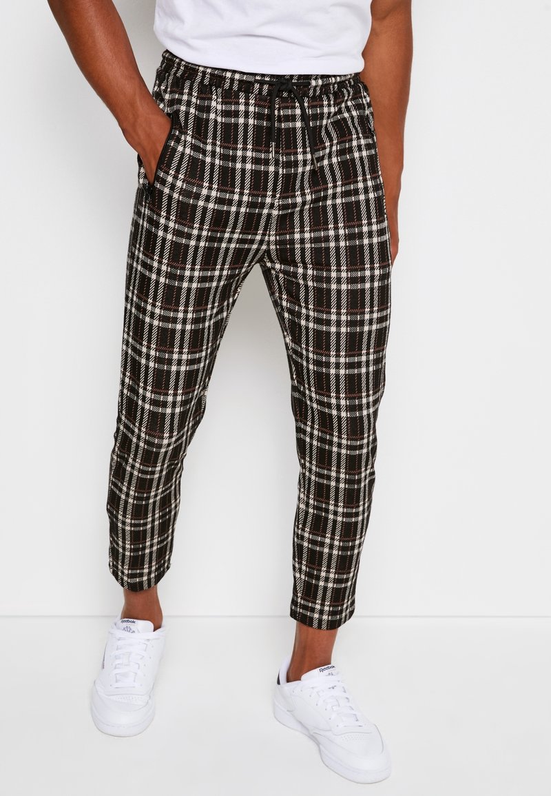 Homme portant un pantalon à carreaux noir et blanc avec une taille élastique et des baskets blanches, debout les mains dans les poches devant un fond blanc.