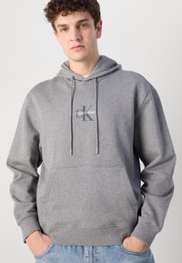 Pilkas medvilnės gobtuvas su priekinėmis kengūros kišenėmis, raišteliu reguliuojamu gobtuvu ir Calvin Klein logotipu ant krūtinės. Su puriomis rankovėmis ir apatine dalimi.