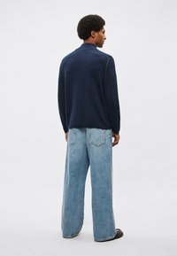 Tengerészkék kötött pulóver magas nyakkal és finom varrási részletekkel, világos színű, széles szárú farmernadrággal párosítva. Egyszerű, minimalista design.