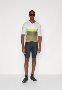 Maglia da ciclismo bianca, oliva e verde neon con maniche corte, zip frontale e tessuto traspirante. Abbinata a pantaloni neri aderenti e scarpe.