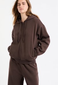 Brązowa bluza z kapturem na zamek, z długimi rękawami i kieszeniami po bokach. Miękka, szczotkowana tkanina z prążkowanymi mankietami i dołem. Swobodny krój.