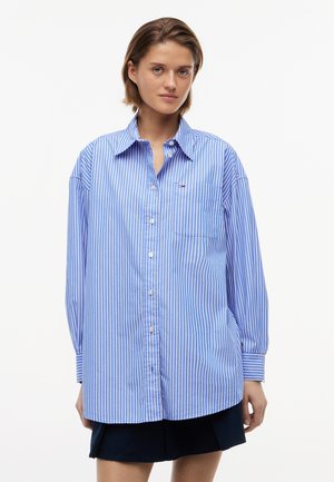 Junge Person trägt ein übergroßes blau-weiß gestreiftes Hemd mit vertikalen Streifen und dunkle Shorts vor weißem Hintergrund.