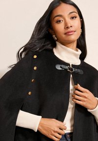 Černý plášť s zlatými knoflíky na rameni, s černým koženým uzávěrem. Převlečený přes smetanový rolák, který ukazuje jemné textury.