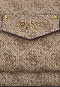 Sac à main en tissu texturé marron avec un motif en diamant, arborant un logo doré "GUESS EST 1981" et des accents dorés sur une bordure foncée.