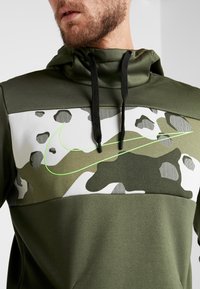 Sudadera color verde oliva con diseño camuflado; presenta patrones en blanco y verde oscuro y un cordón negro. Acento del logo de Nike en verde neón.