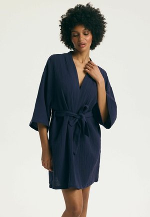 Žena s kučeravými vlasmi, oblečená v tmavomodrom textúrovanom župane dĺžky ku kolenám, zaviazanom v páse, stojaca pred jednoliatym pozadím.