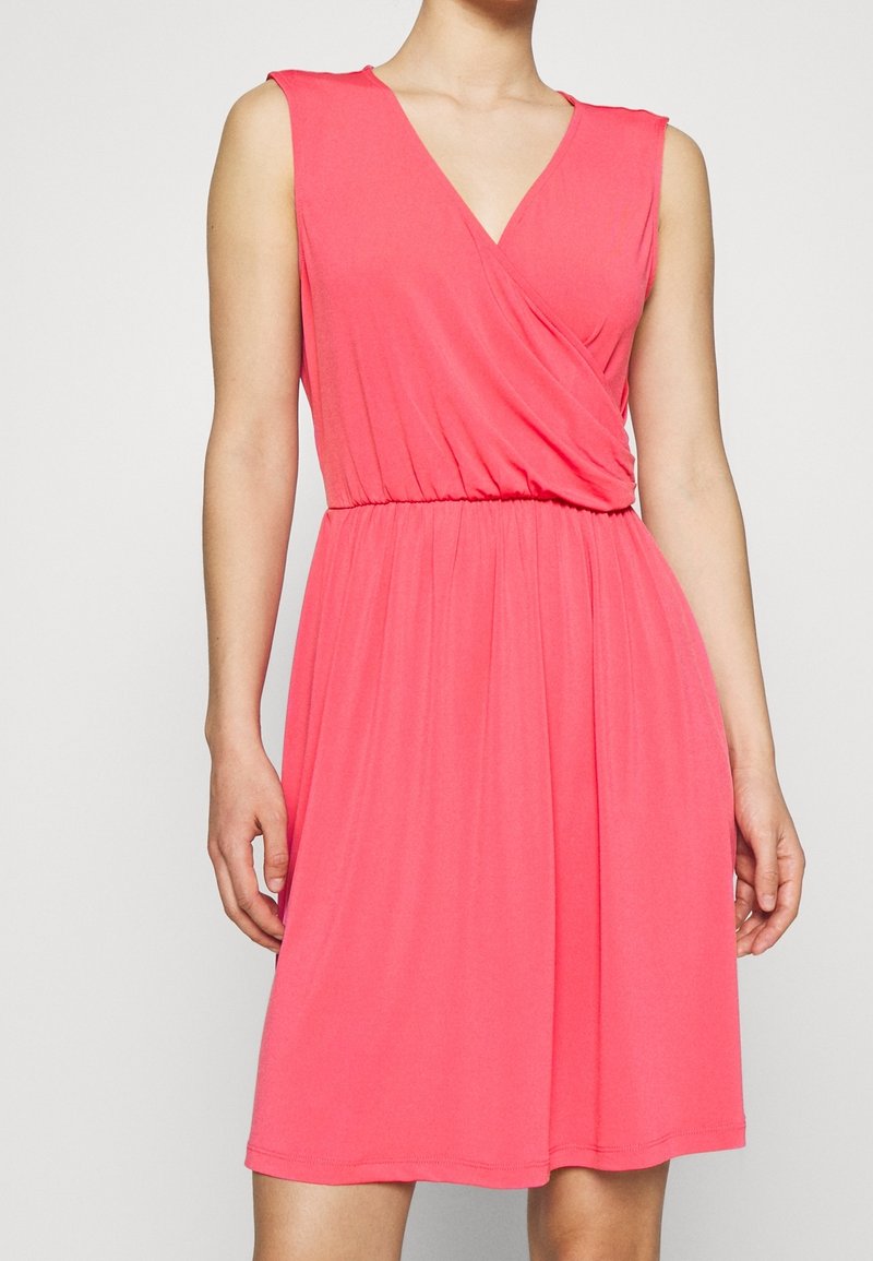 Robe corail rose sans manches avec un corsage cache-cœur à encolure en V et une taille élastique, jupe longueur genou, portée par une personne debout.